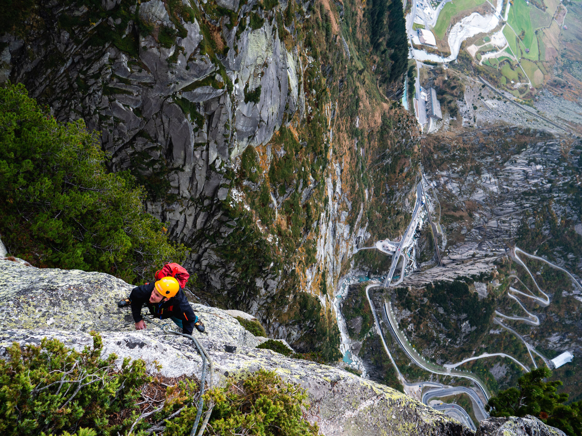 A climber ascending an exposed multipitch route, with a switchback road visible far below in the valley